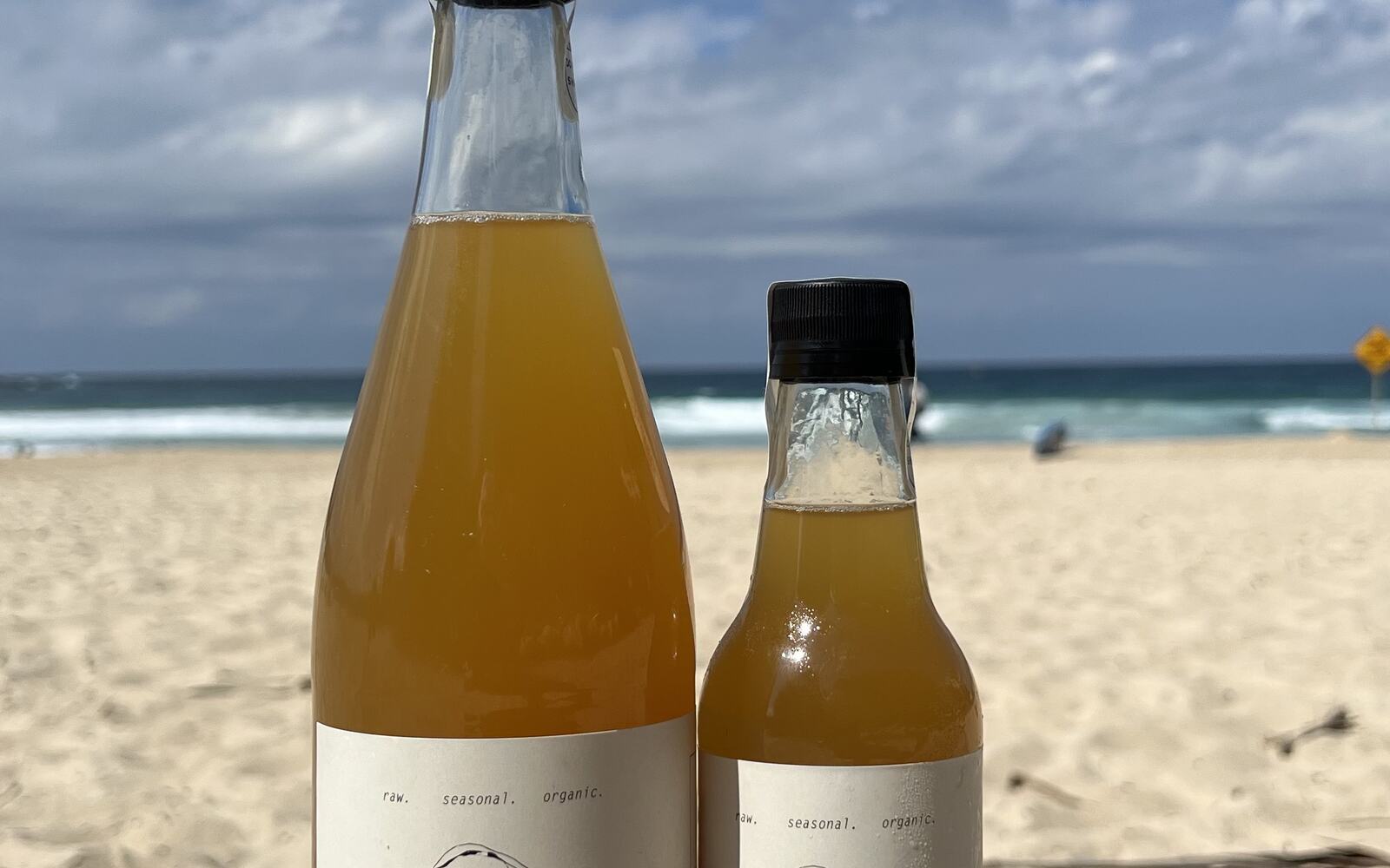 Both bottle sizes of Marjorie Cultures standing on sand at the beach.